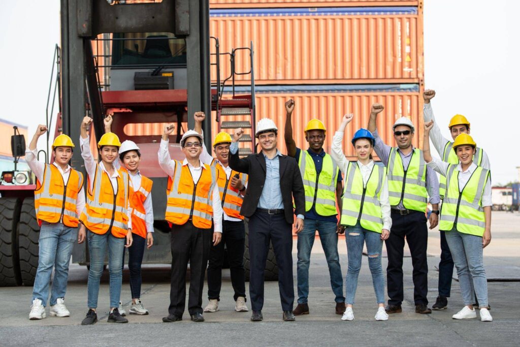 A diverse group of happy construction workers and engineers celebrating in front of a large yellow crawler bulldozer at a sunny construction site, representing successful project completion and teamwork.
